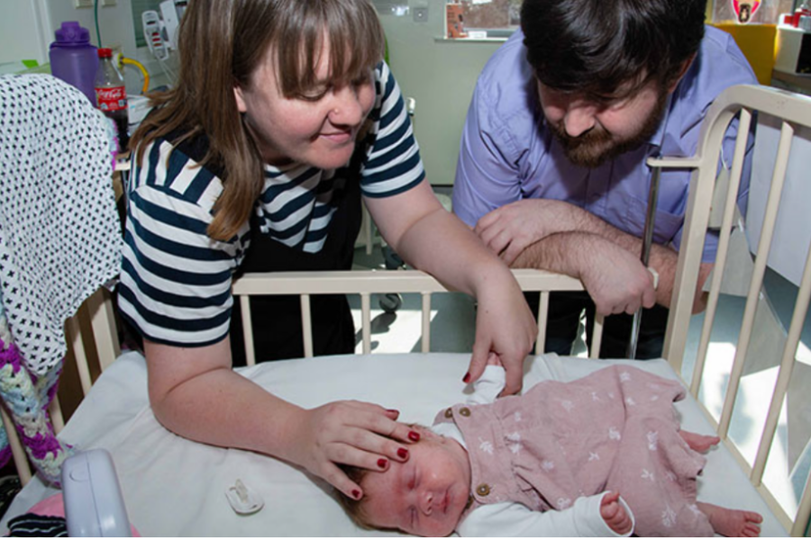 A woman and a man lean over a hospital crib, gently interacting with a newborn baby lying on its back. The baby is dressed in a pink outfit and is resting, while the woman softly touches the baby’s head. The setting is a hospital room, with medical equipment and supplies visible in the background.